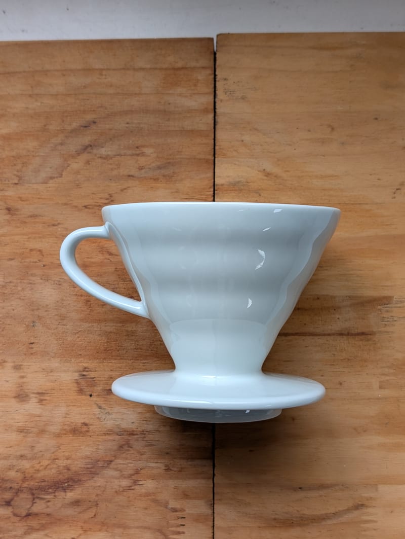 Hario V60 cone dripper in white ceramic with its handle and flared base, standing on a wooden board